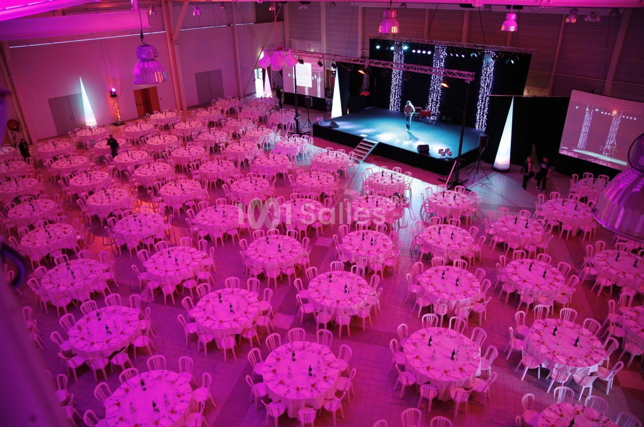 Salle de réception illuminée en rose avec de nombreuses tables rondes dressées et une scène au fond.