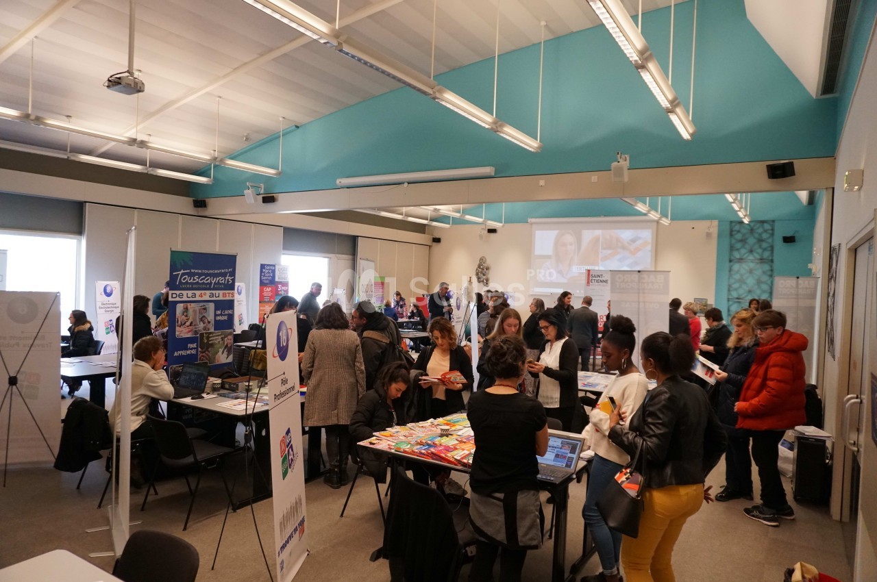 Salle animée avec des stands d'information, des visiteurs échangeant et un écran projetant une présentation.