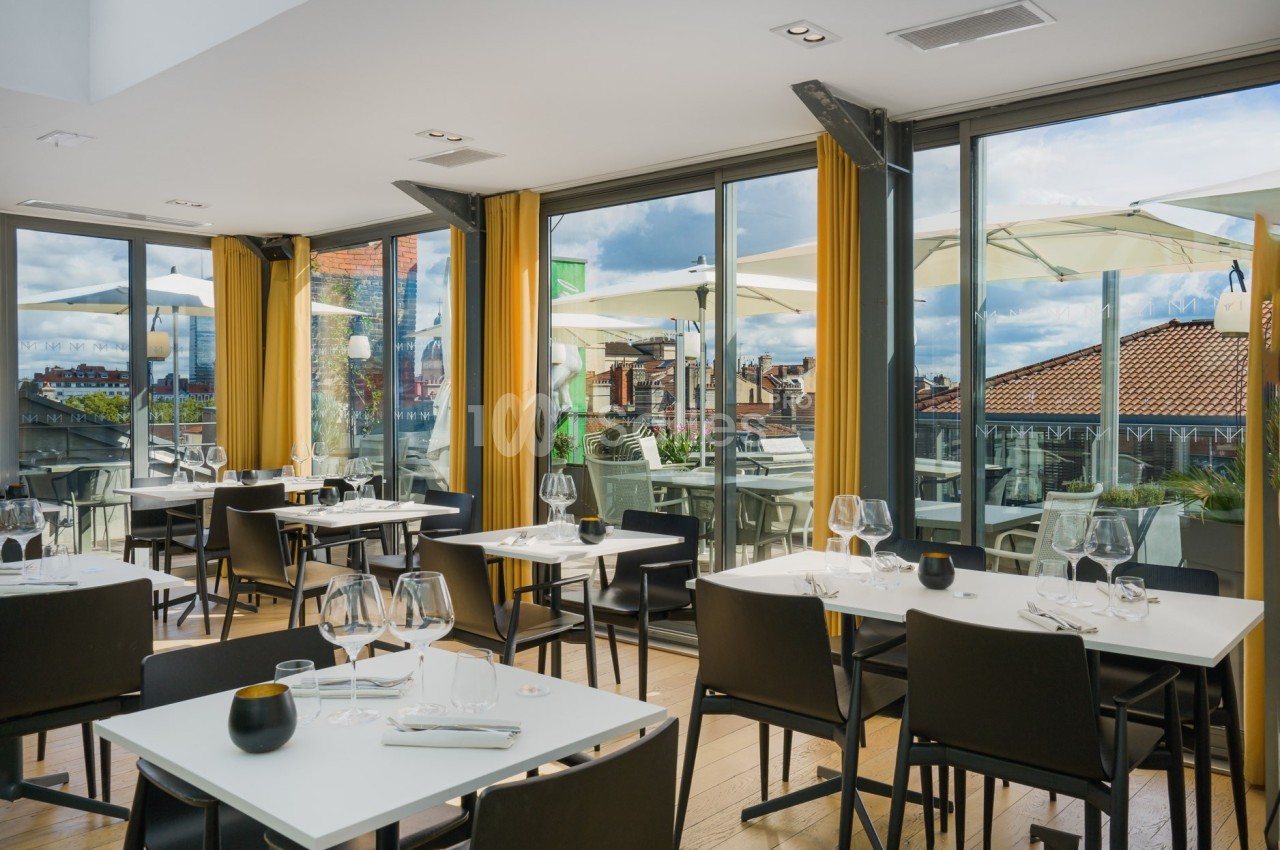 Salle de restaurant lumineuse avec grandes baies vitrées donnant sur une terrasse équipée de parasols.