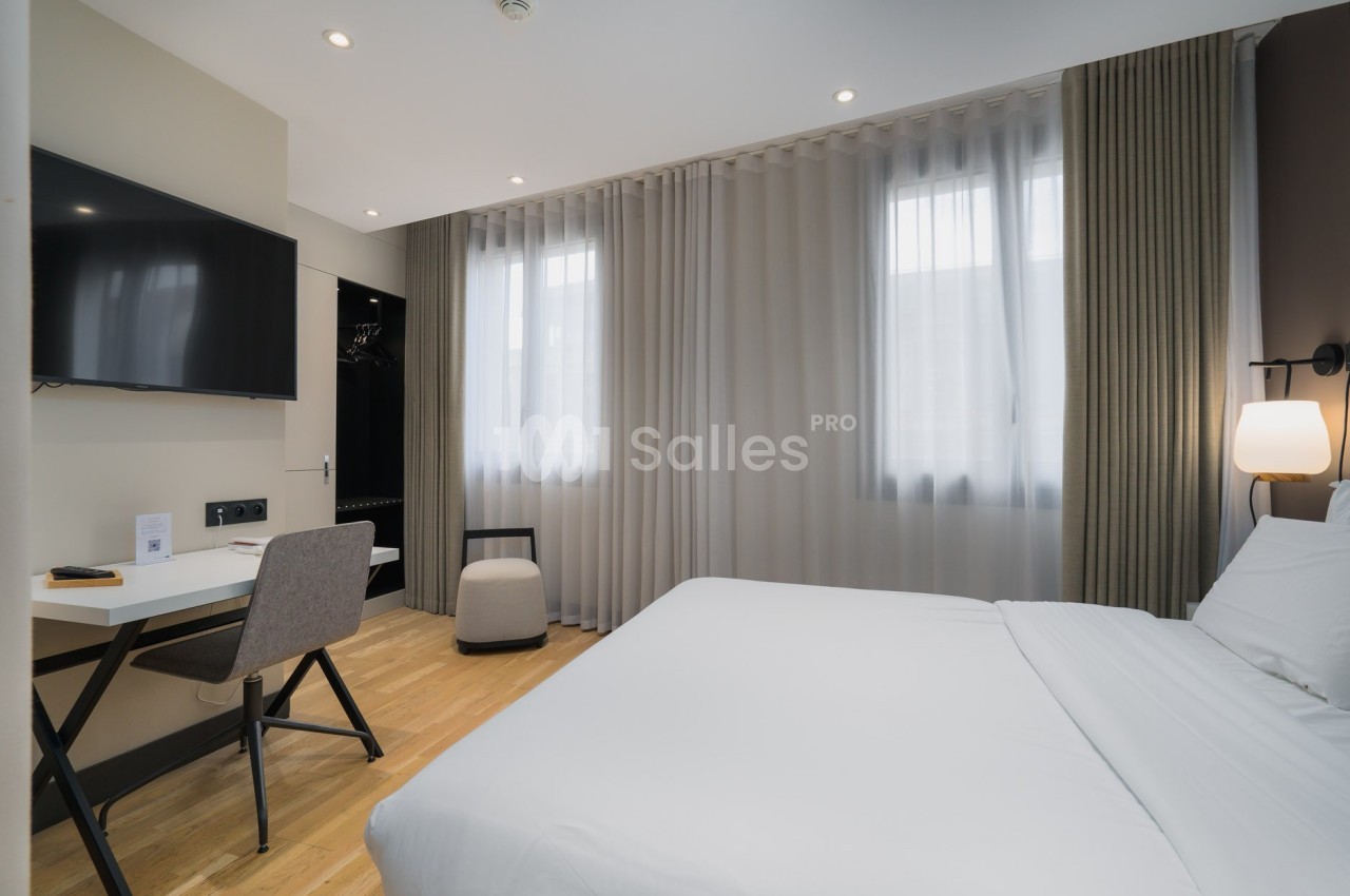 Chambre lumineuse avec lit double, bureau, chaise, télévision murale et rideaux translucides devant les fenêtres.