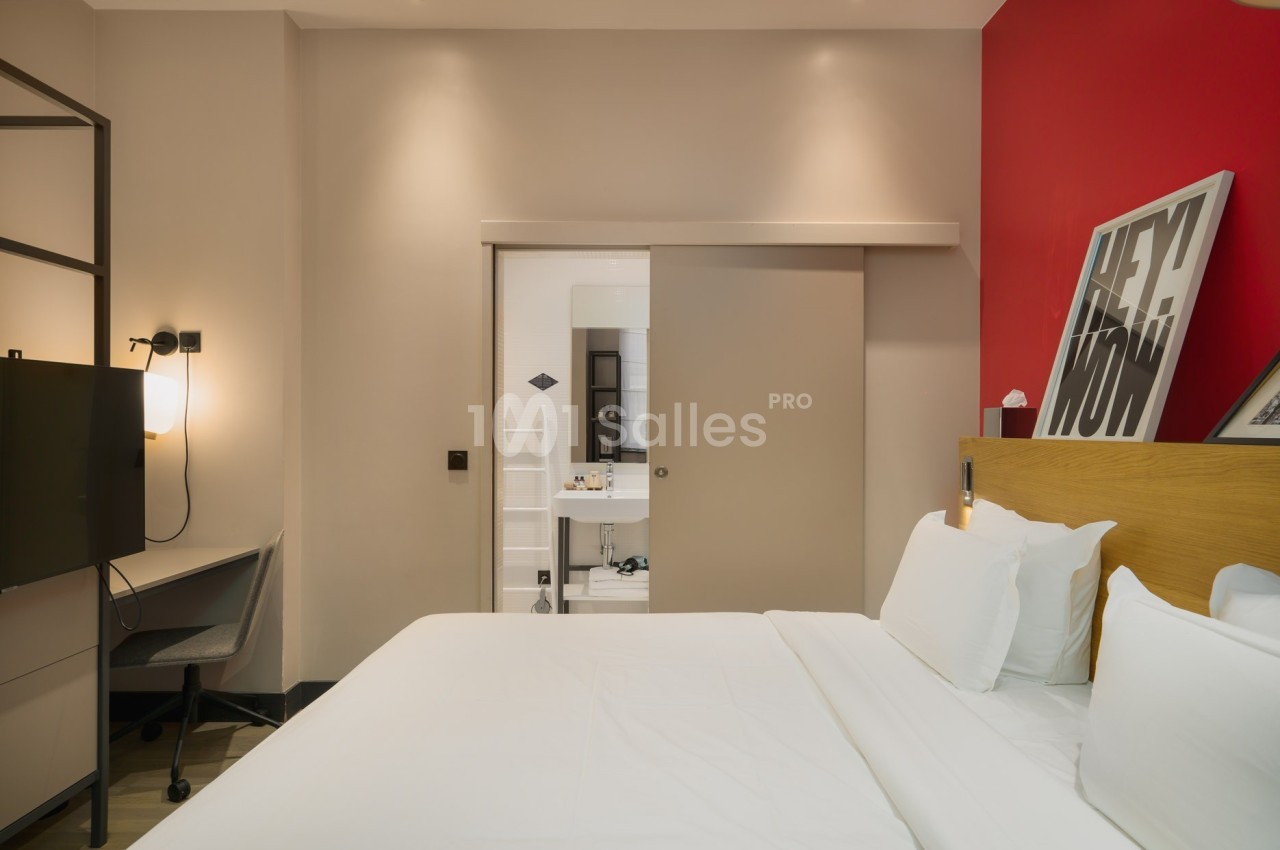 Chambre d'hôtel moderne avec lit double, bureau, télévision, et salle de bain visible à travers une porte coulissante.