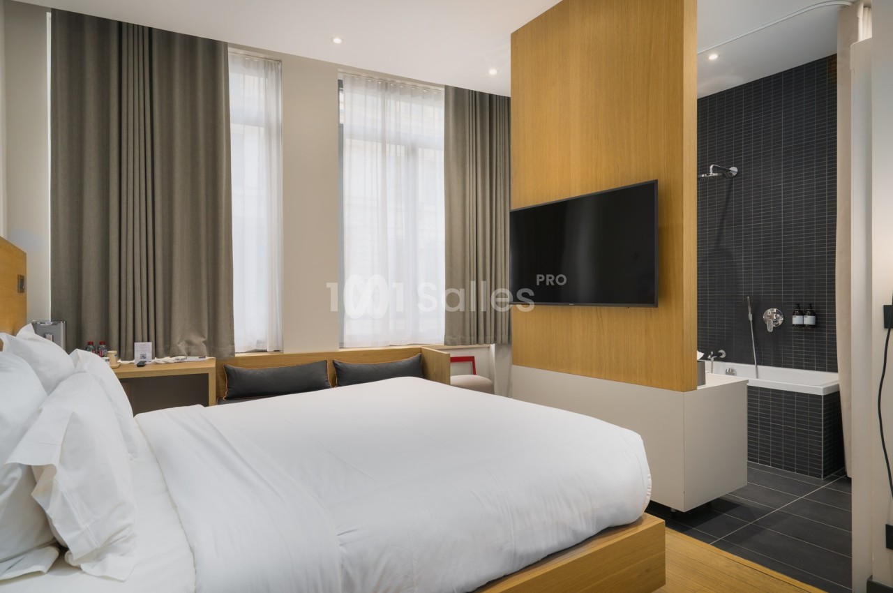 Chambre d'hôtel moderne avec lit double, télévision murale, grande fenêtre et salle de bain attenante avec baignoire.
