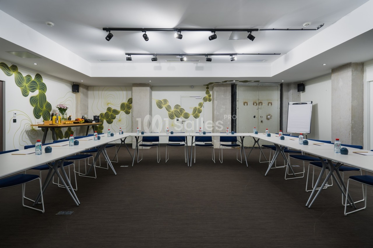 Salle de réunion lumineuse avec tables disposées en U, chaises bleues, tableau de conférence et buffet en arrière-plan.