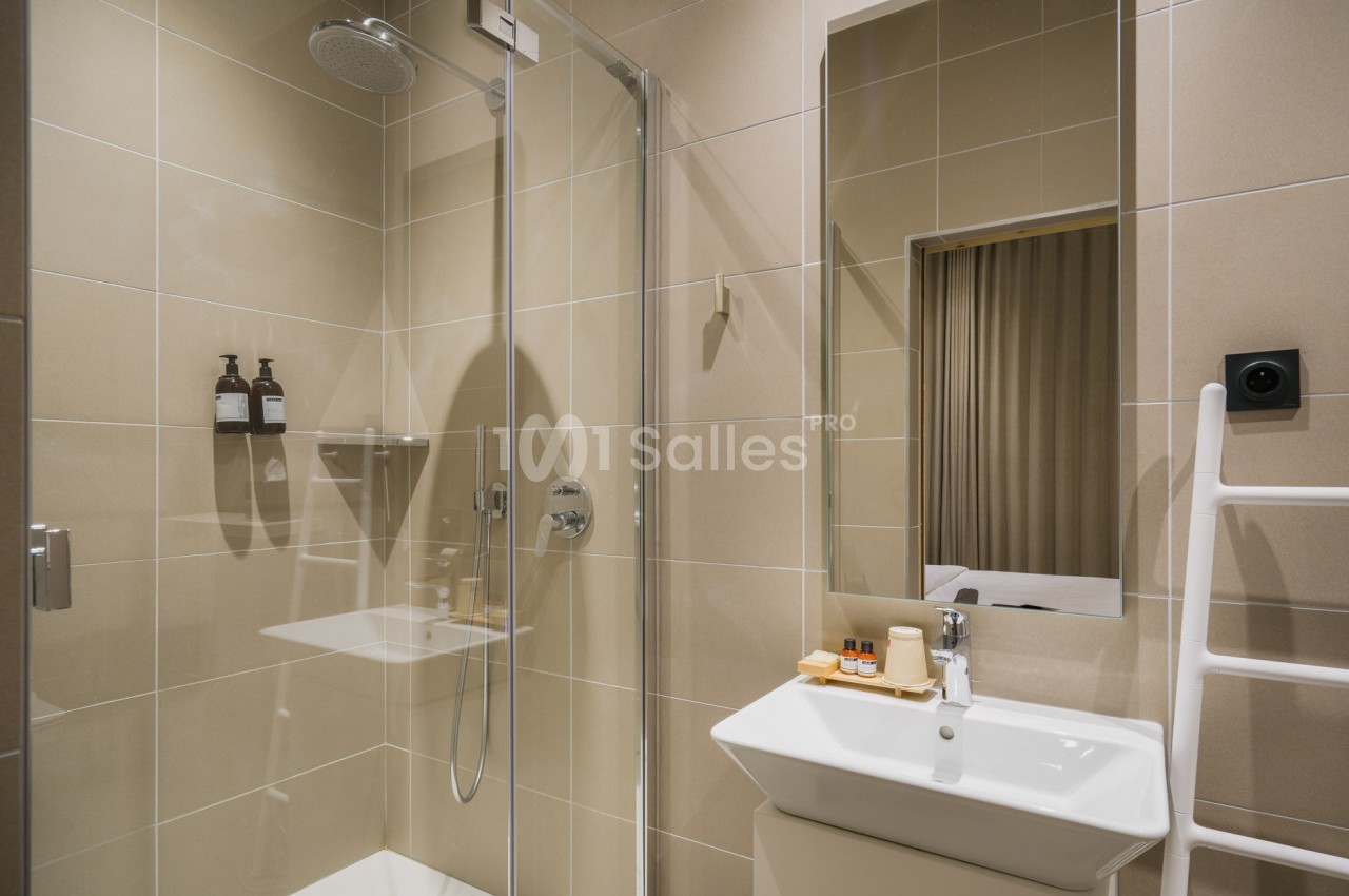 Salle de bain moderne avec douche vitrée, lavabo blanc, miroir, et accessoires de toilette sur un mur carrelé beige.