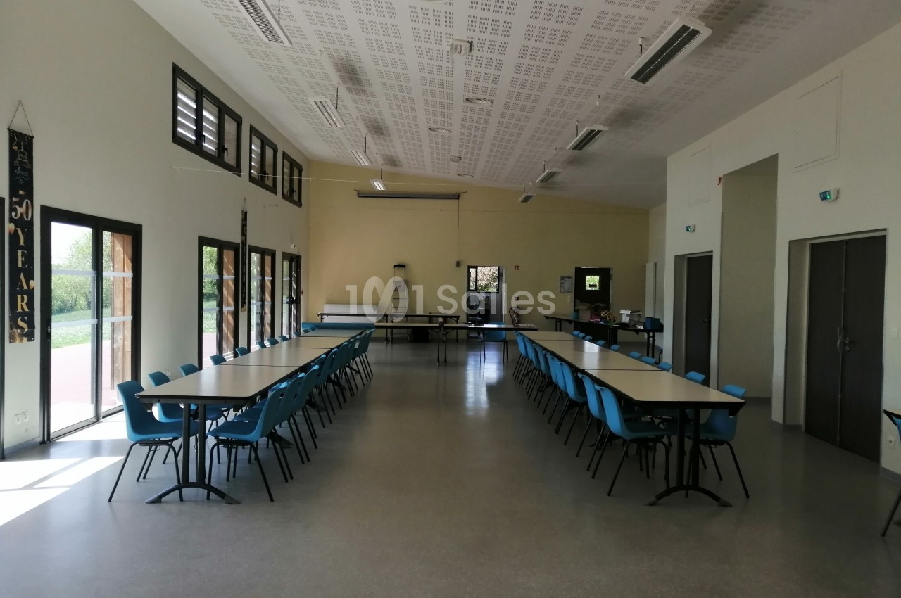 Salle lumineuse avec deux rangées de tables et chaises bleues, grandes fenêtres et portes vitrées sur les côtés.