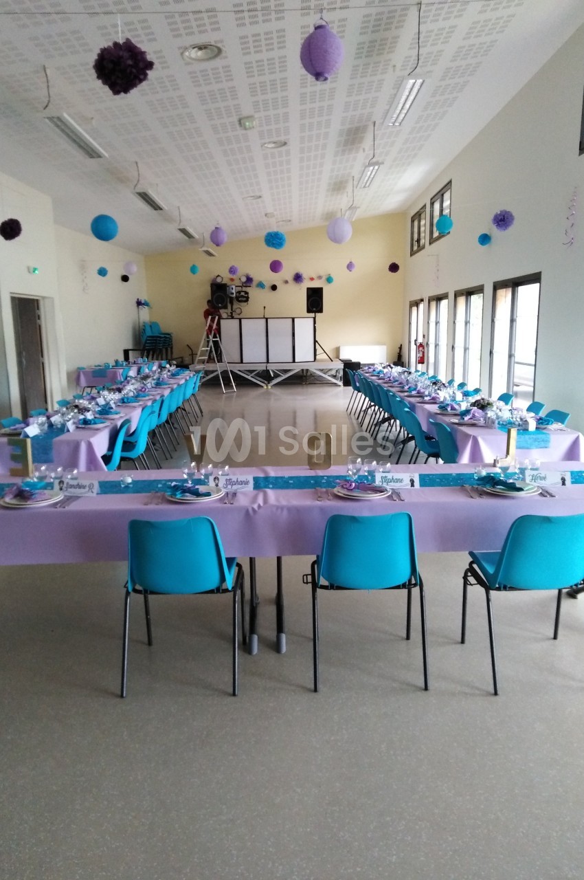 Salle décorée pour un événement avec tables dressées en violet et bleu, chaises alignées et espace pour une piste de danse.