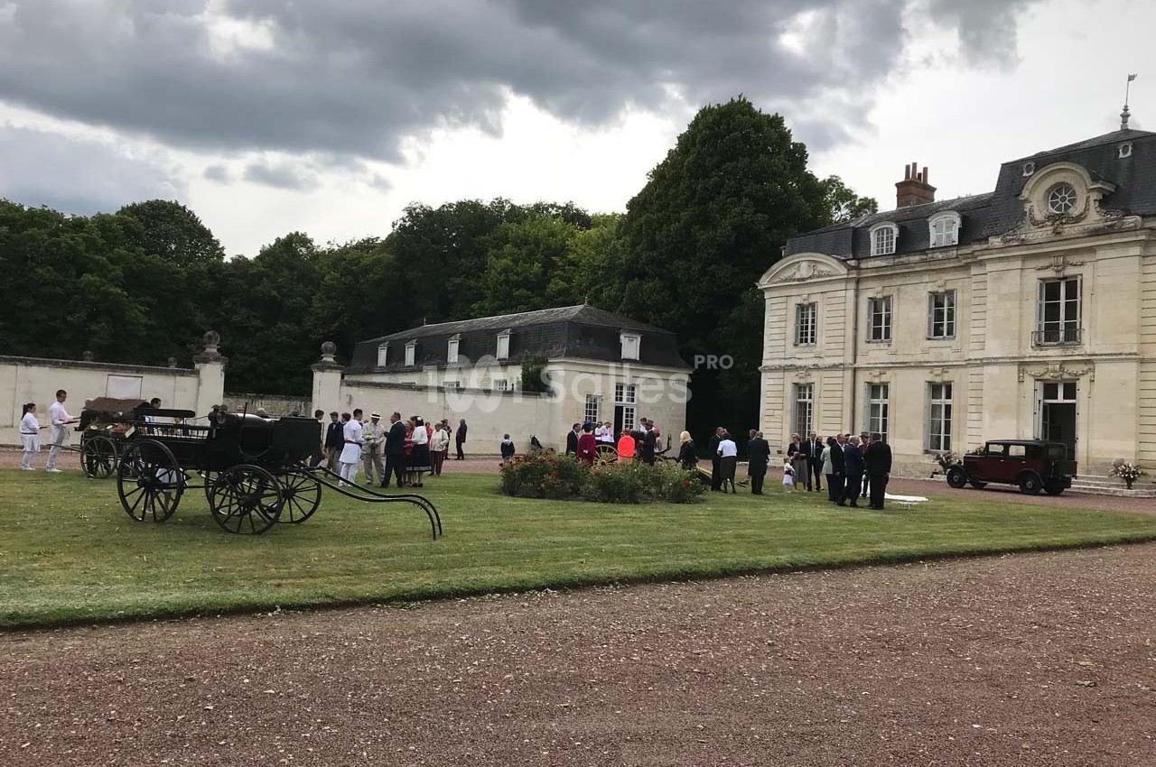 Groupe de personnes rassemblées dans la cour d'un château entouré de verdure, avec des calèches en premier plan.