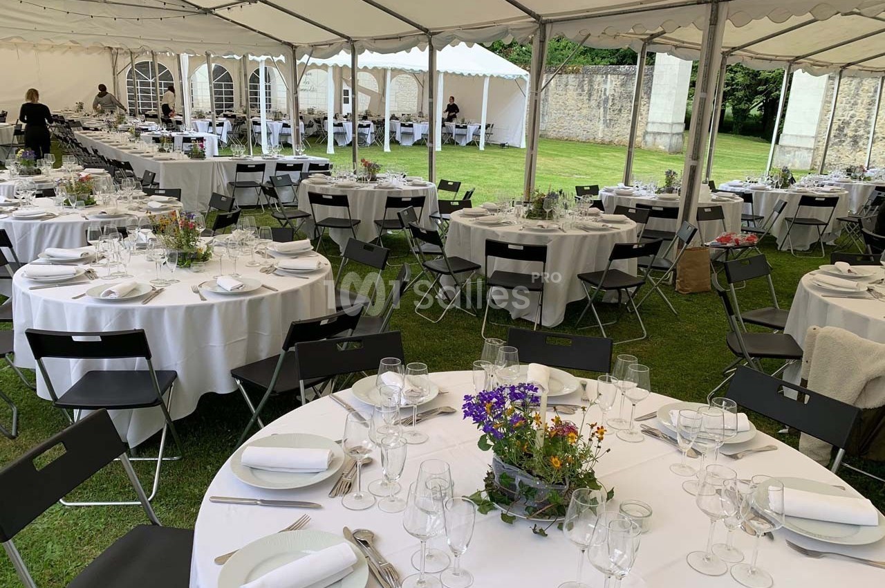Tables rondes dressées avec nappes blanches, couverts et décorations florales sous une grande tente en extérieur.