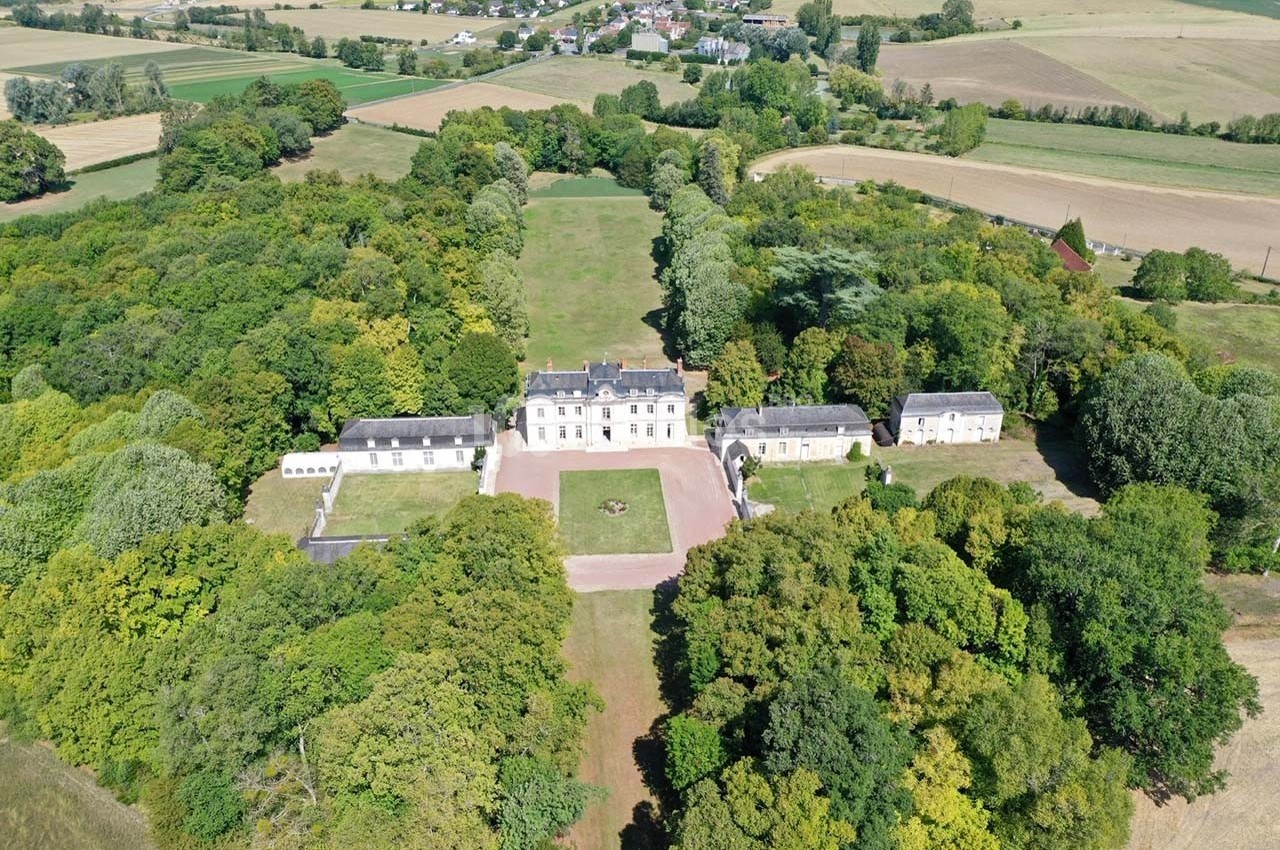 Vue aérienne d'un château entouré de jardins et de forêts, avec un village visible à l'arrière-plan.