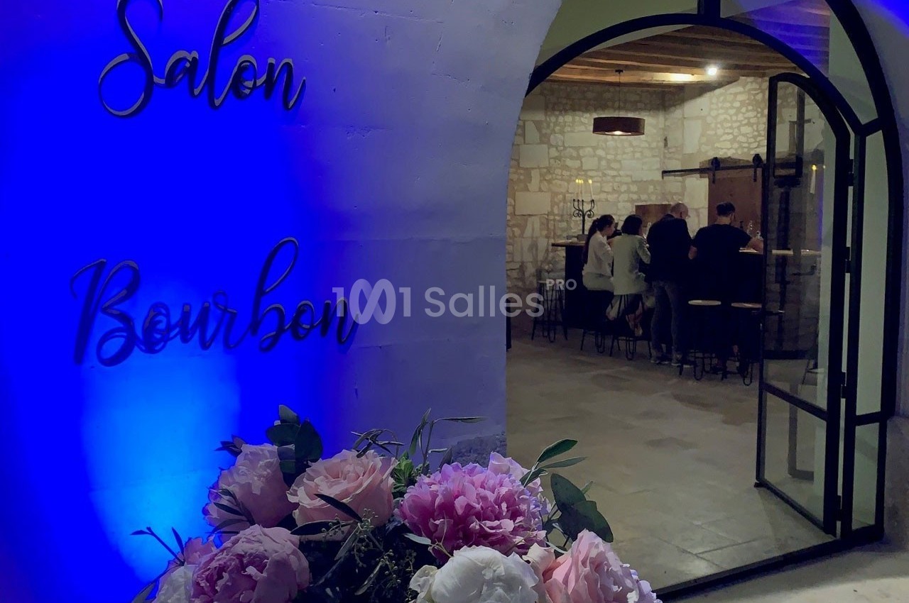 Entrée d'un salon éclairé en bleu, avec des fleurs au premier plan et des personnes discutant autour d'un bar à l'intérieur.