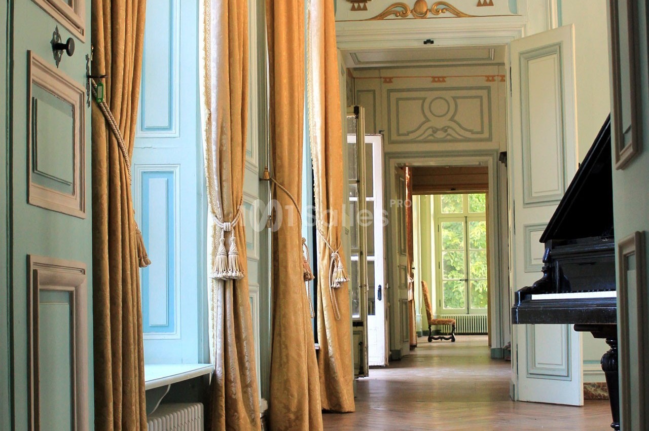 Intérieur lumineux avec des rideaux dorés, un piano noir et des portes ouvertes donnant sur un couloir élégant.