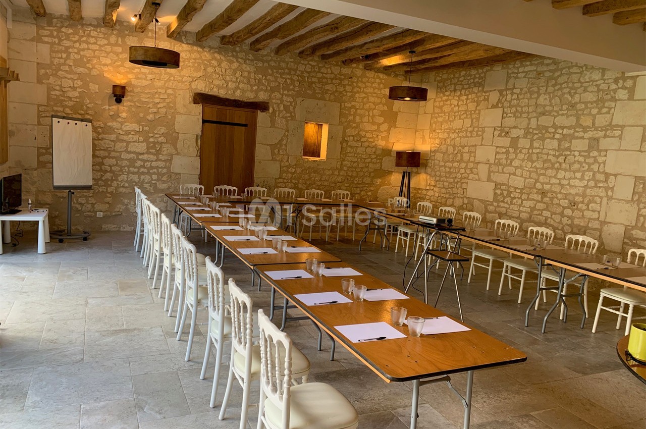 Salle de réunion lumineuse avec murs en pierre, tables disposées en U et chaises blanches alignées.