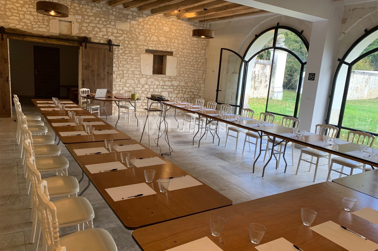 Salle lumineuse aménagée en configuration de réunion avec tables alignées, chaises blanches et grandes baies vitrées.