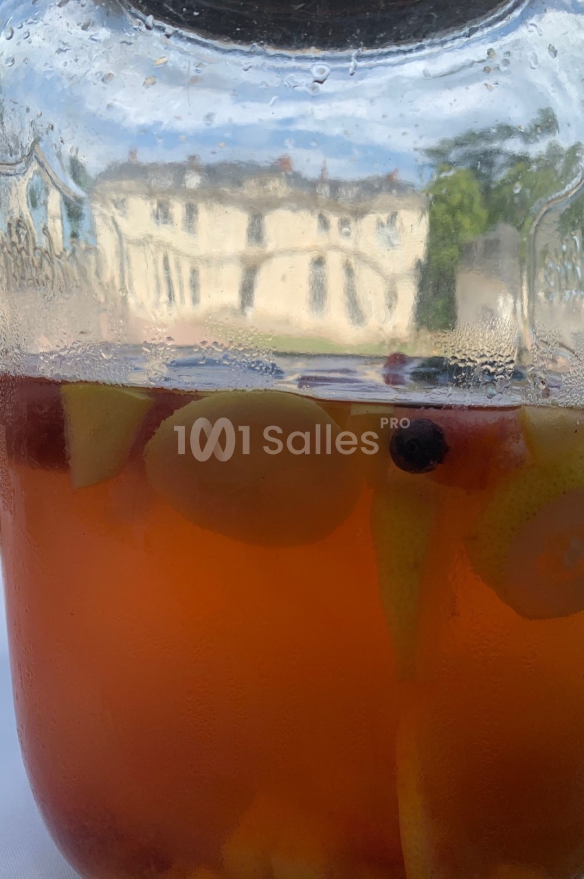 Bocal en verre contenant une boisson fruitée avec des morceaux de fruits, posé devant un bâtiment flou en arrière-plan.