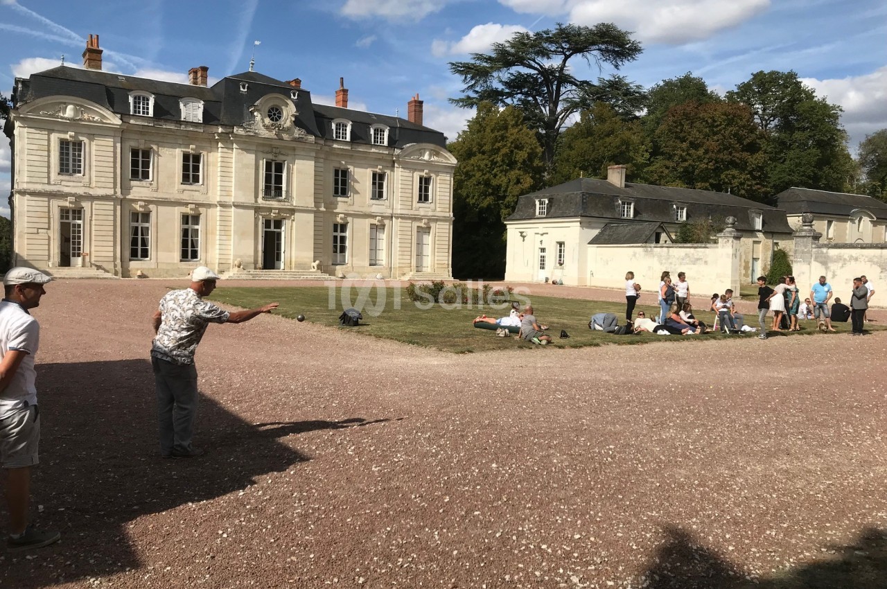 Un groupe de personnes joue à la pétanque devant un bâtiment ancien entouré de verdure.