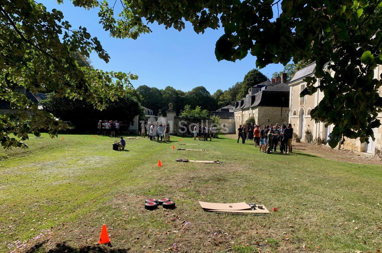 Groupe de personnes rassemblées dans une cour herbeuse près de bâtiments anciens, avec des activités au sol.