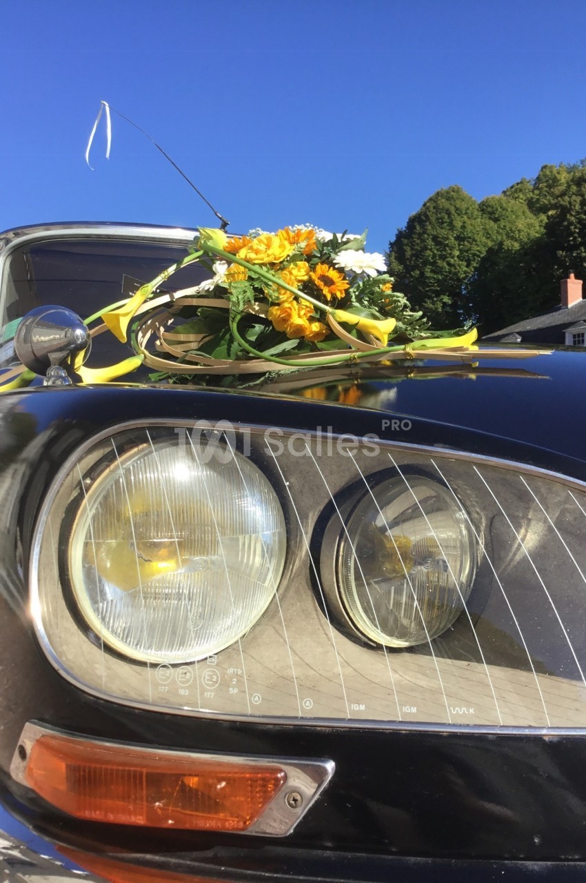Bouquet de fleurs jaunes et blanches posé sur le capot d'une voiture ancienne noire, sous un ciel bleu.