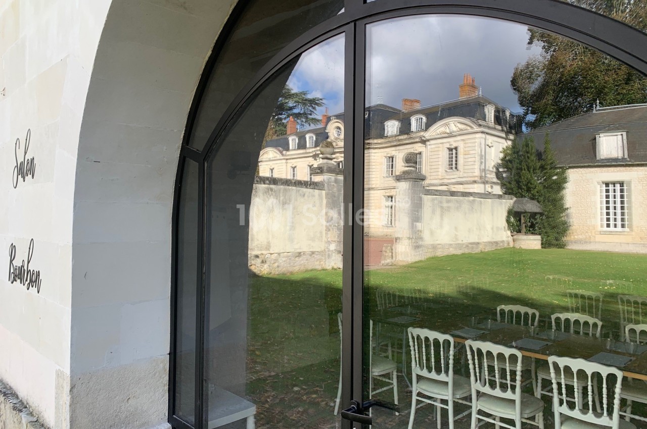 Reflet d'un château dans une baie vitrée, avec des chaises blanches disposées à l'intérieur près d'une table.