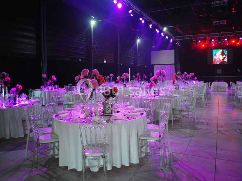 Salle de réception décorée avec des tables rondes, nappes blanches, chaises transparentes et éclairage coloré.