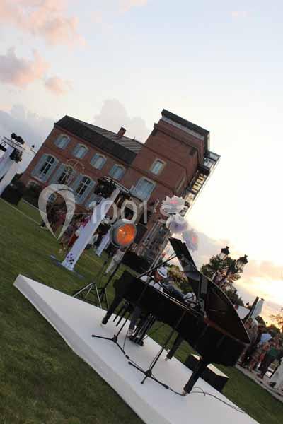 Un piano à queue noir sur une scène extérieure devant un bâtiment en briques, avec des spectateurs en arrière-plan.