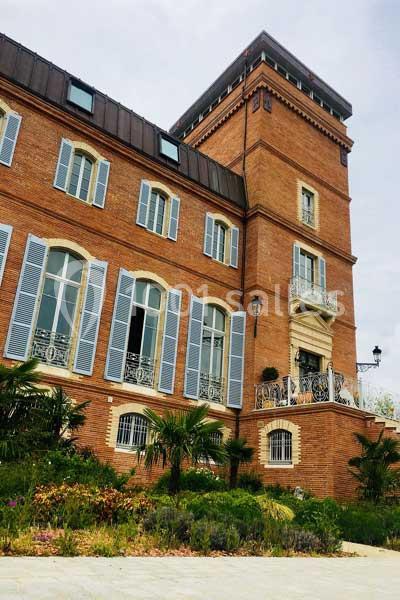 Bâtiment en briques rouges avec volets bleus, une tour et un jardin fleuri au premier plan.