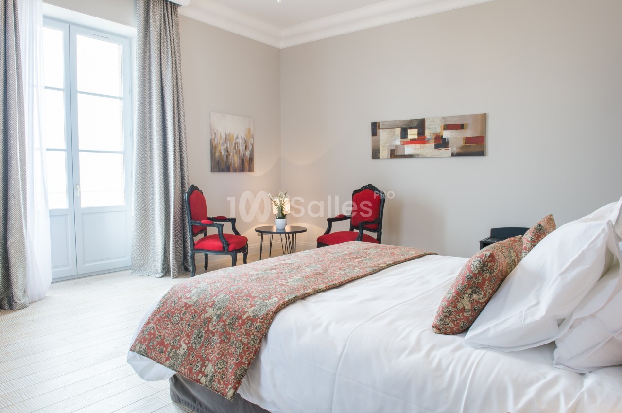 Chambre lumineuse avec lit double, deux fauteuils rouges, table basse, tableau mural et grande fenêtre.