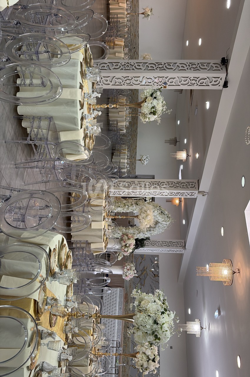 Salle de réception élégante avec tables rondes dressées en ivoire, chaises transparentes, colonnes décoratives blanches et arrangements floraux blancs et dorés.