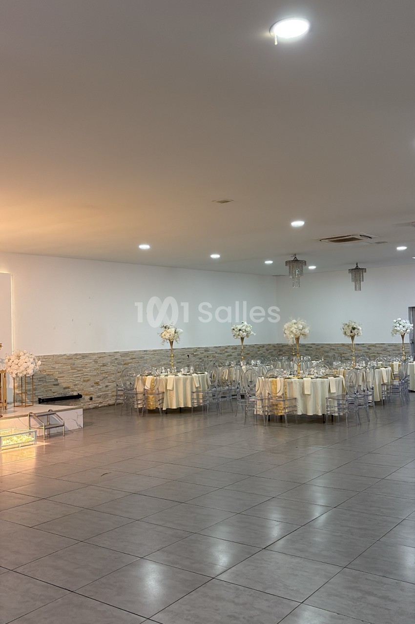 Salle de réception aménagée avec tables rondes dressées, chaises transparentes et bouquets de fleurs blanches.