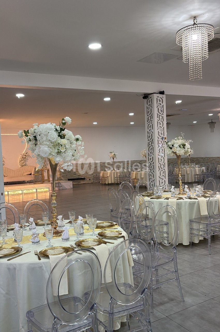 Salle de réception dressée avec tables rondes nappe blanche, chaises transparentes et décoration florale blanche.