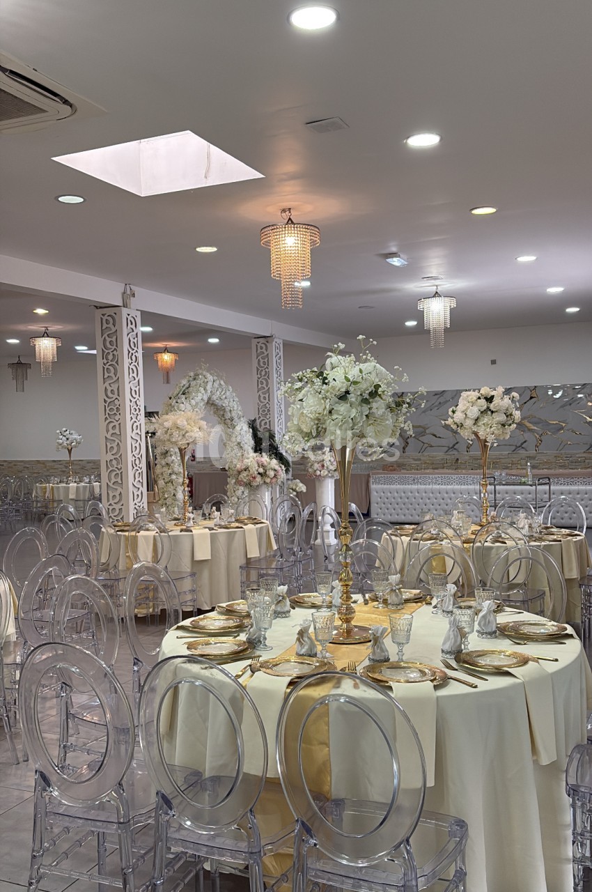 Salle de réception dressée avec tables rondes en nappes crème, chaises transparentes et compositions florales blanches et champagne.