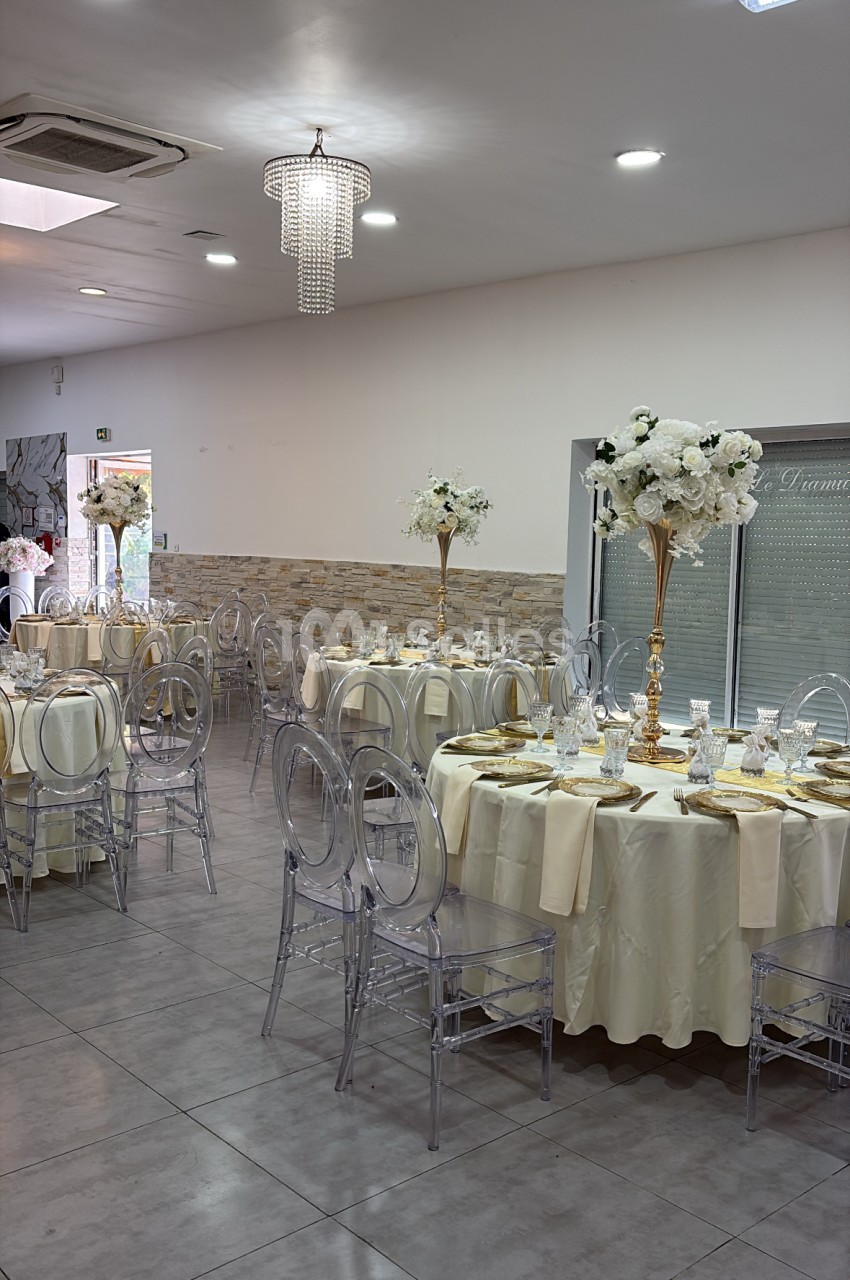 Salle intérieure dressée pour réception - tables rondes beige, chaises transparentes, lustres cristal, fleurs blanches.
