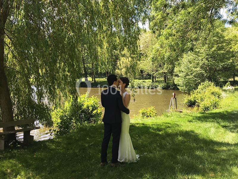 Un couple en tenue de mariage se tient près d'un étang entouré de verdure sous un arbre.