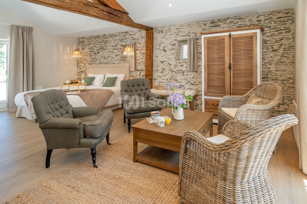 Chambre lumineuse avec murs en pierre, lit double, fauteuils, table basse et armoire en bois dans un décor rustique.