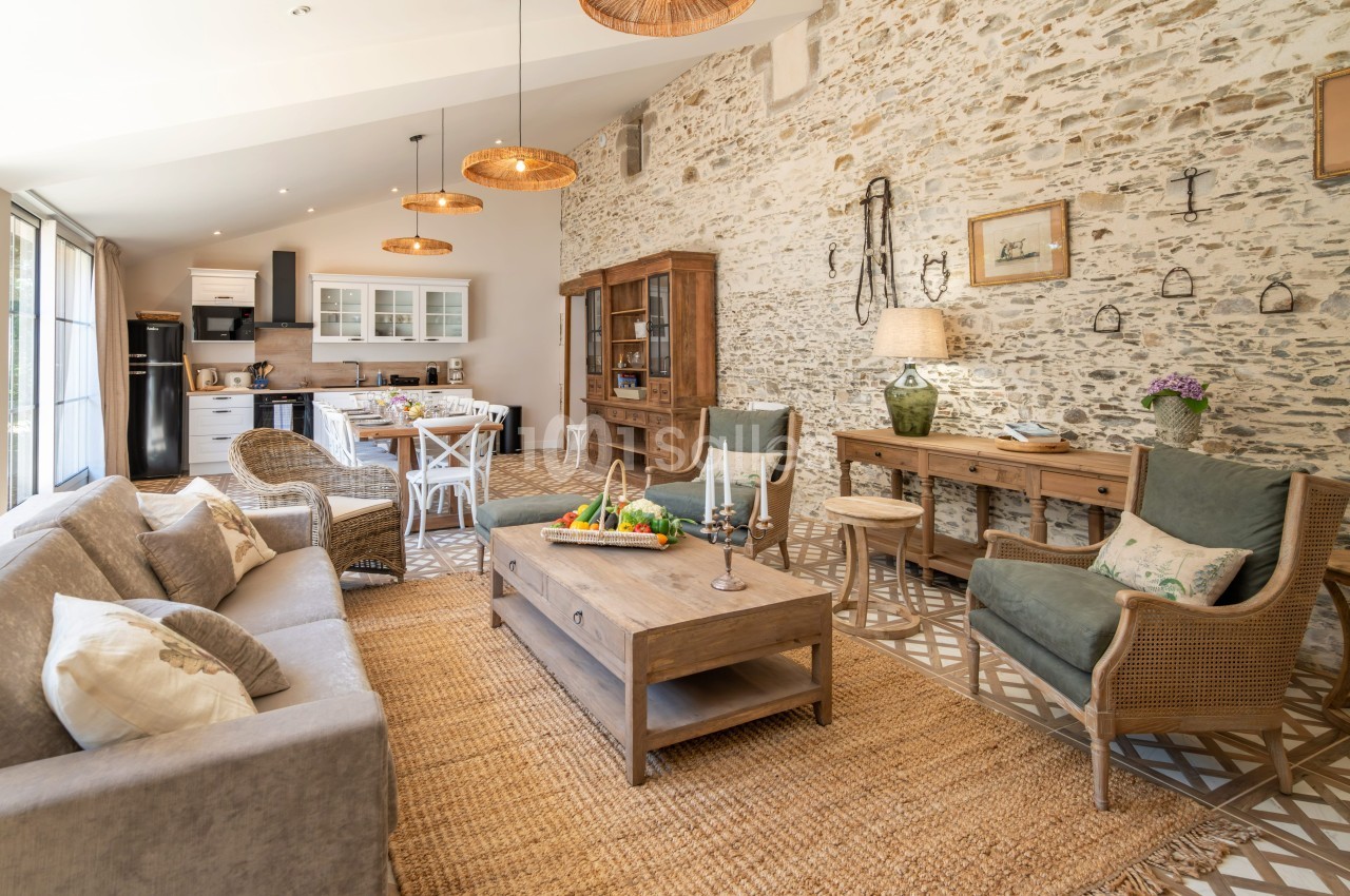 Salon lumineux avec murs en pierre, mobilier en bois, canapé beige, et cuisine ouverte en arrière-plan.