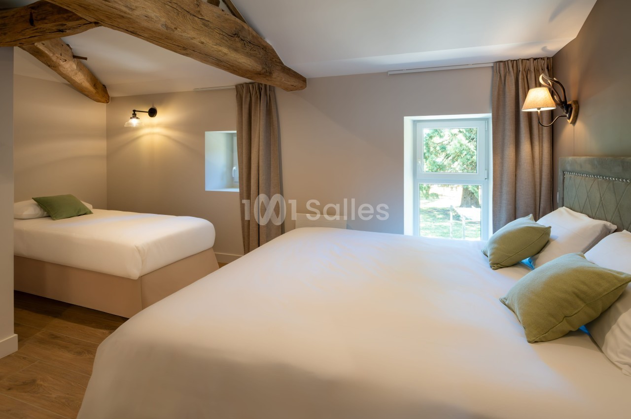Chambre lumineuse avec deux lits, poutres apparentes, murs clairs, rideaux beige et vue sur un jardin par la fenêtre.