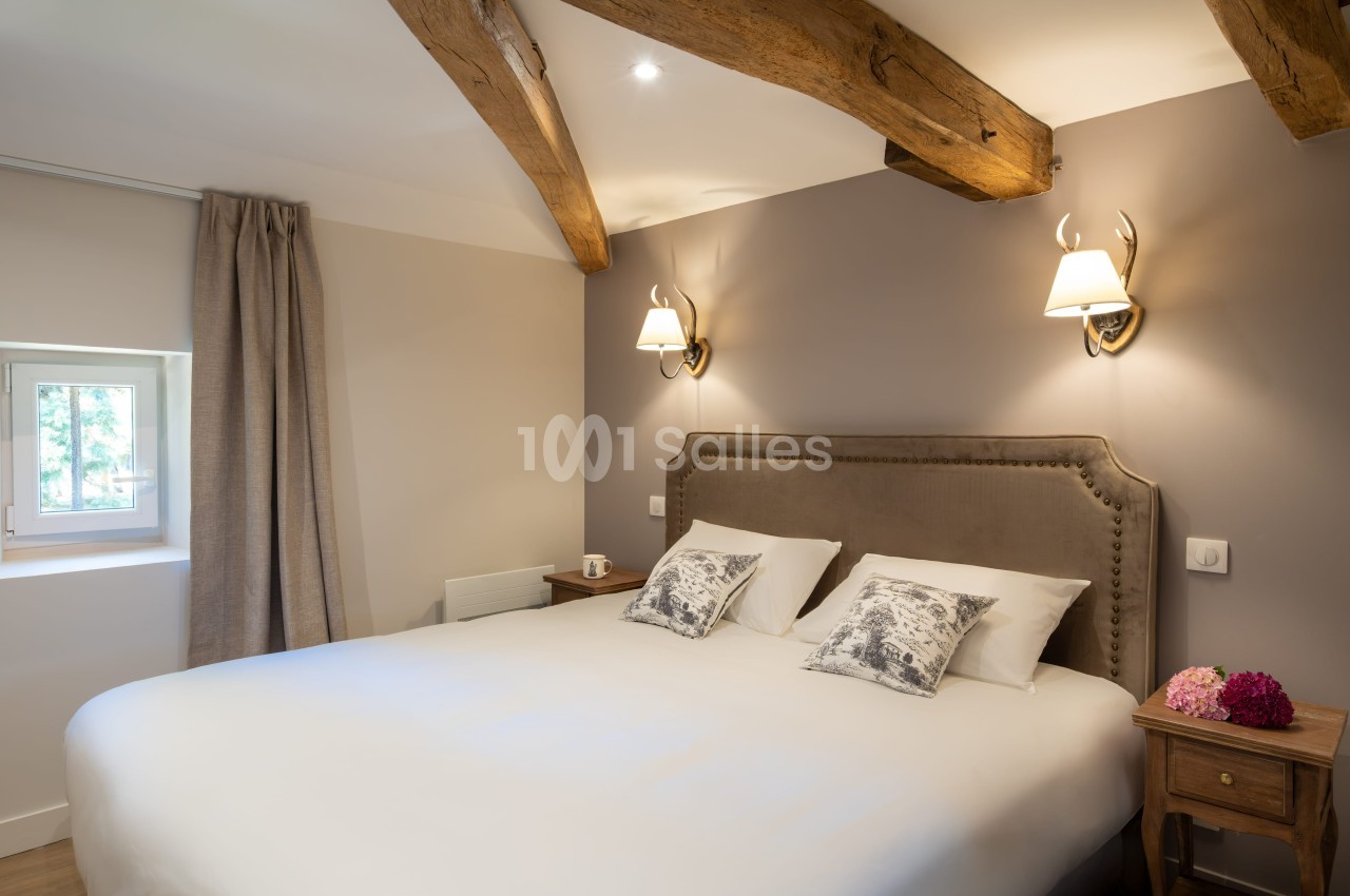 Chambre lumineuse avec lit double, poutres apparentes, murs clairs, lampes murales et table de chevet en bois.