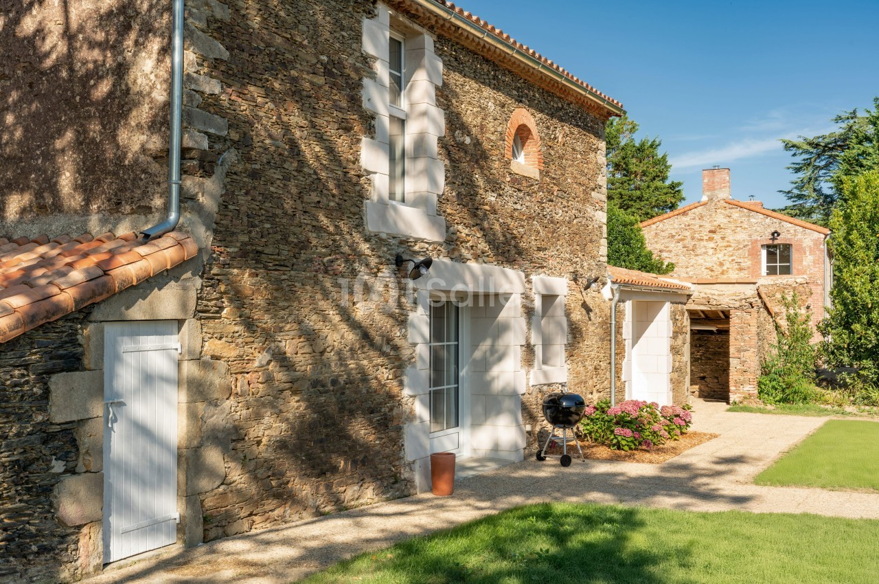 Façade en pierre d'une maison avec portes blanches, barbecue et jardin fleuri sous un ciel bleu.