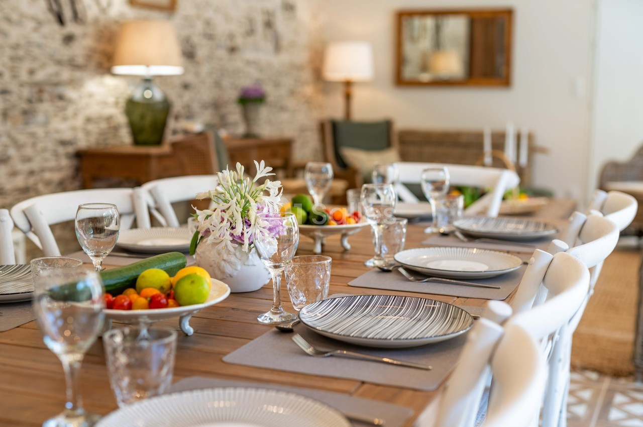 Table en bois dressée avec vaisselle, verres, fruits et fleurs, dans une salle lumineuse au décor chaleureux.