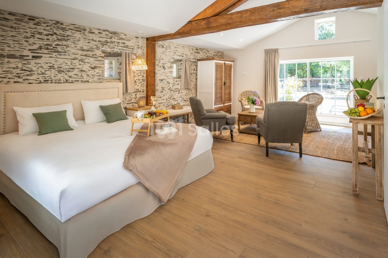 Chambre lumineuse avec lit double, fauteuils, table basse, murs en pierre apparente et grande baie vitrée donnant sur un…
