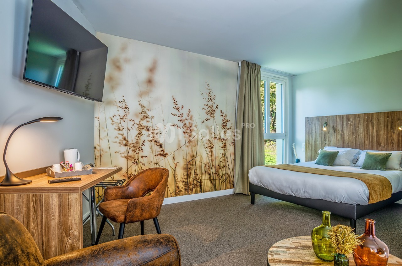 Chambre d'hôtel lumineuse avec lit double, bureau, fauteuils, télévision murale et vue sur un espace vert.