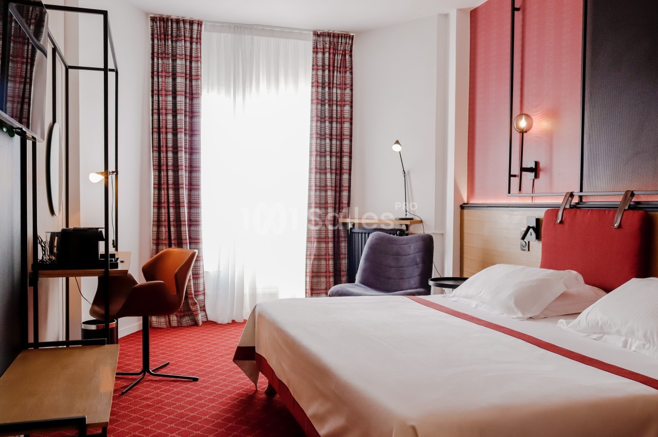 Chambre d'hôtel lumineuse avec un lit double, un bureau, un fauteuil et des rideaux à motifs rouges devant une grande…