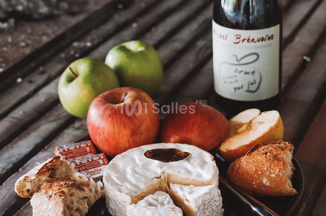 Plateau avec camembert, pommes, pain, beurre et bouteille de cidre sur une table rustique.