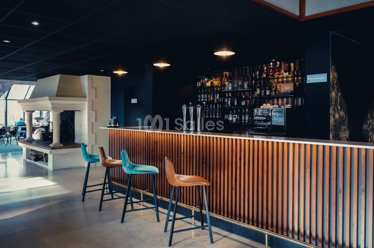 Bar moderne avec comptoir en bois, tabourets colorés et étagères remplies de bouteilles, dans un espace lumineux.