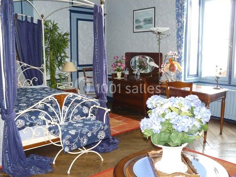 Chambre décorée avec un lit à baldaquin, meubles en bois, fleurs et lumière naturelle entrant par une fenêtre.