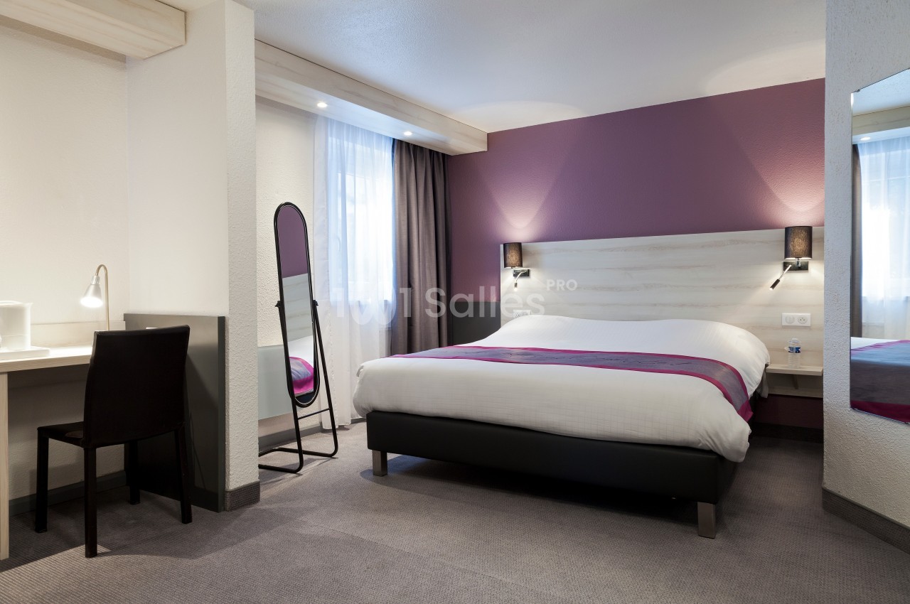 Chambre d'hôtel avec lit double, bureau, chaise, miroir sur pied et éclairage naturel provenant d'une fenêtre.