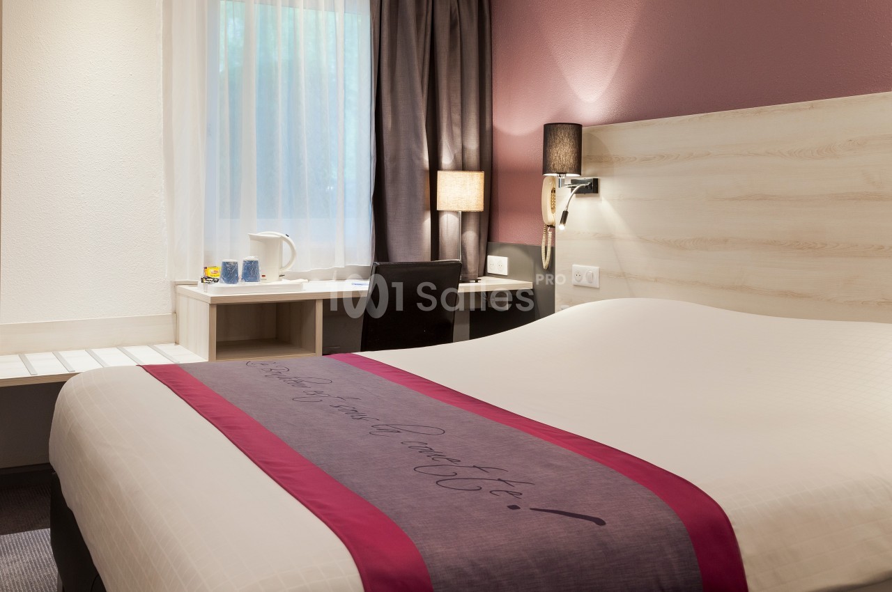 Chambre d'hôtel avec lit double, bureau, chaise, lampe de chevet et fenêtre avec rideaux semi-ouverts.
