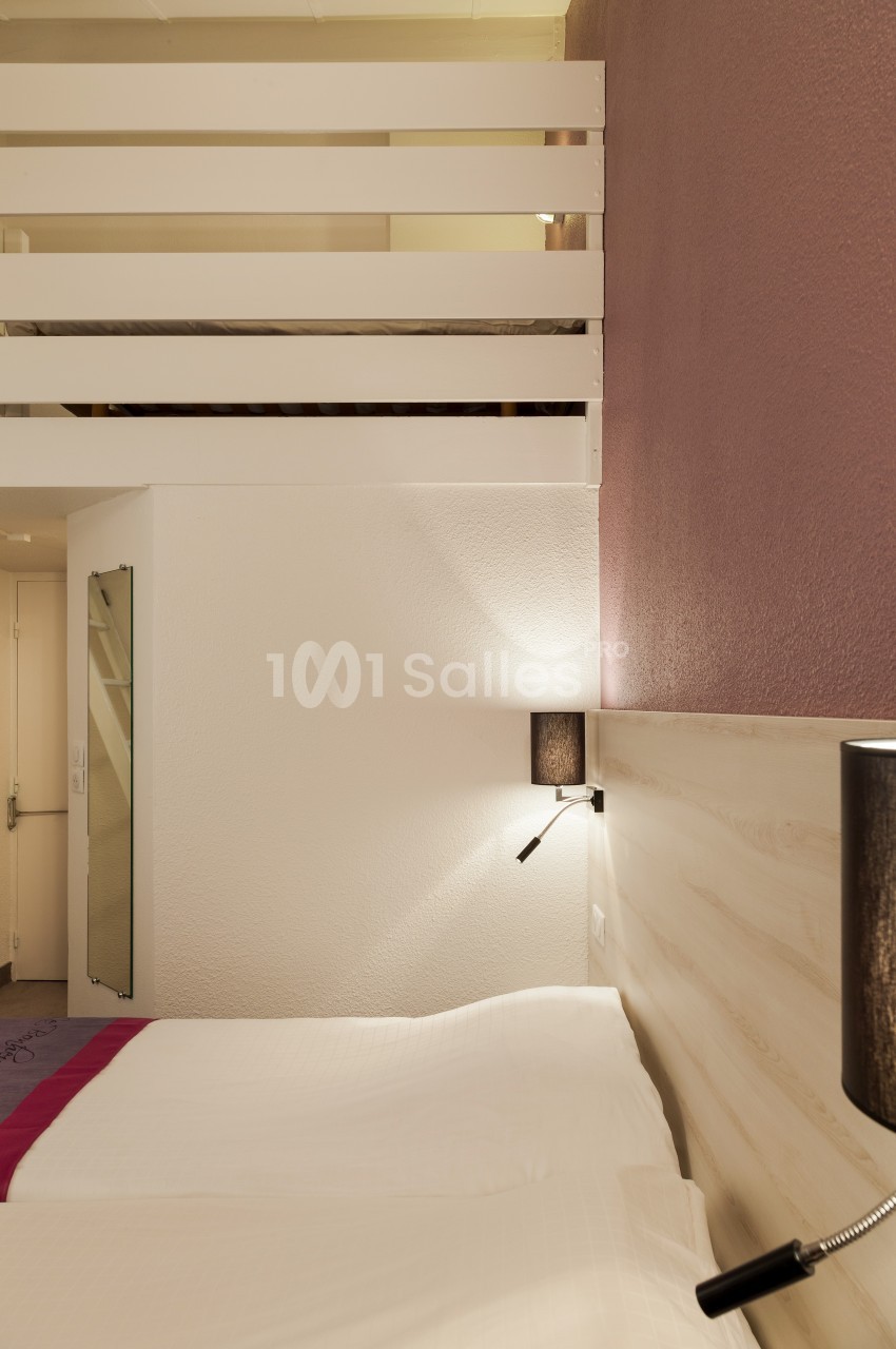 Lit double avec tête de lit en bois clair, éclairage mural et mezzanine visible dans une chambre aux tons neutres.