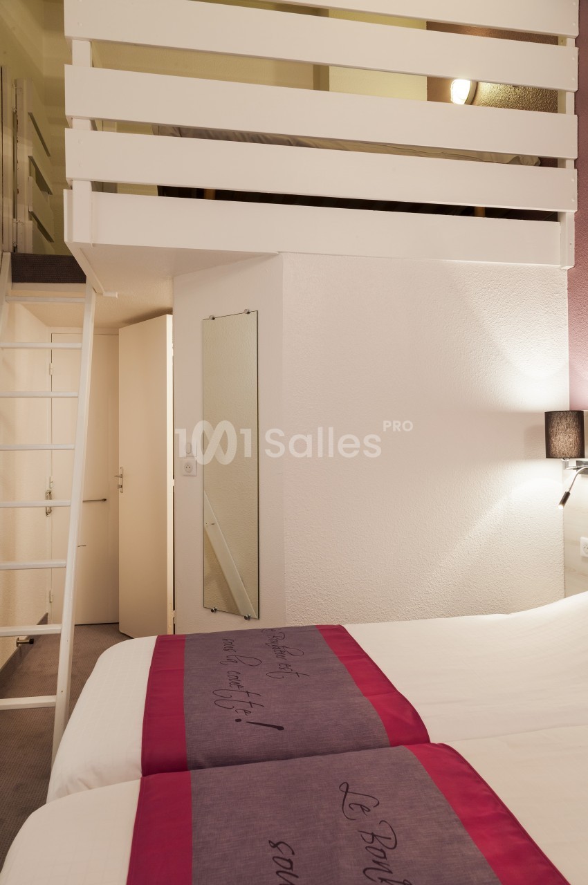 Chambre avec mezzanine, lit double au premier plan, échelle menant à l'étage supérieur, éclairage doux.