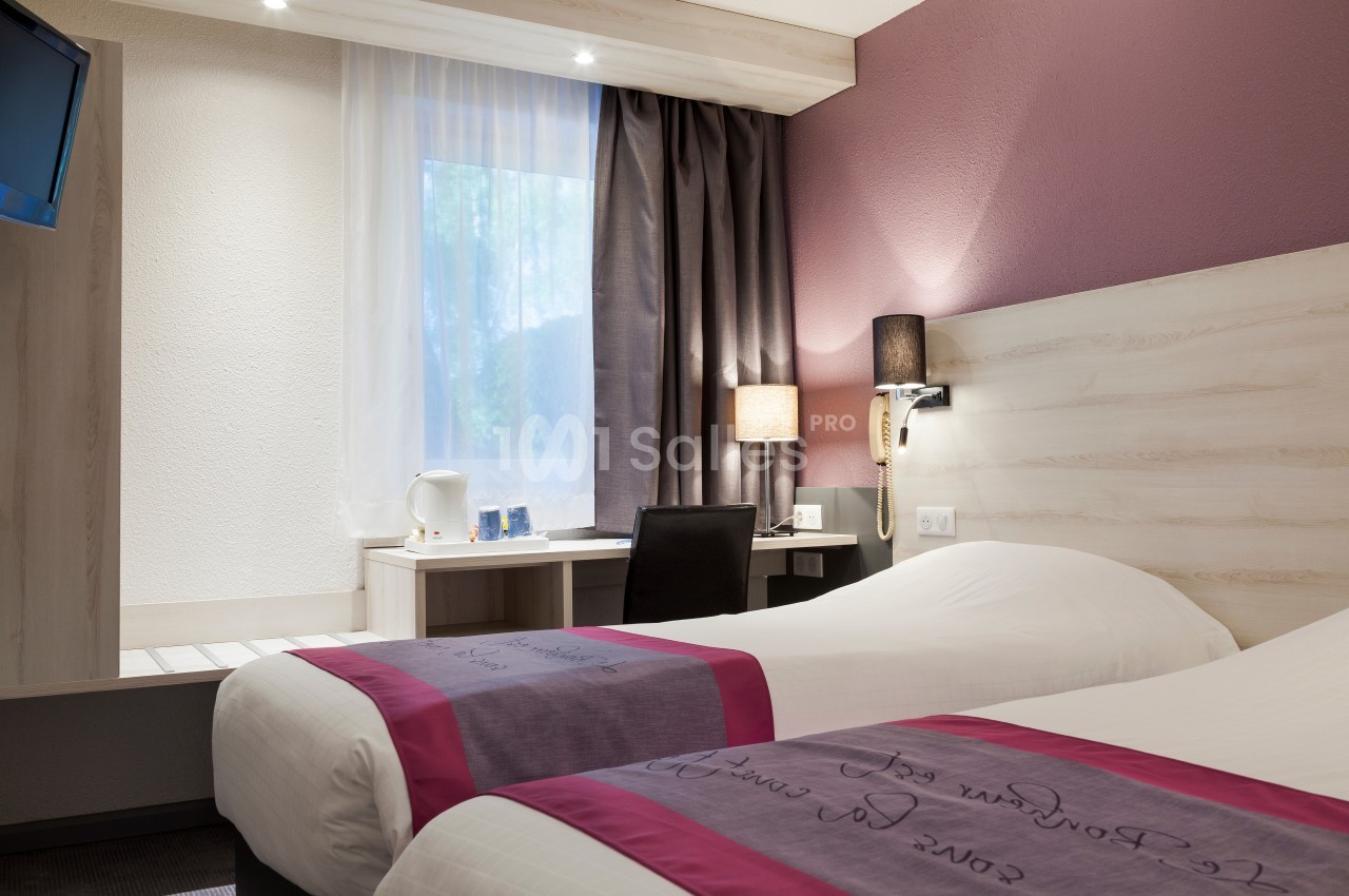 Chambre d'hôtel avec deux lits simples, bureau équipé, lampe de chevet et fenêtre avec rideaux.