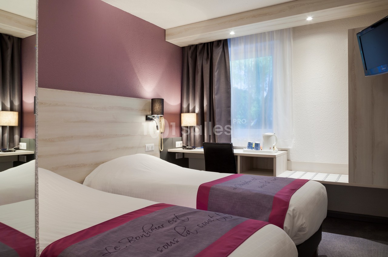 Chambre d'hôtel avec deux lits simples, bureau, télévision murale, miroir et fenêtre avec rideaux.